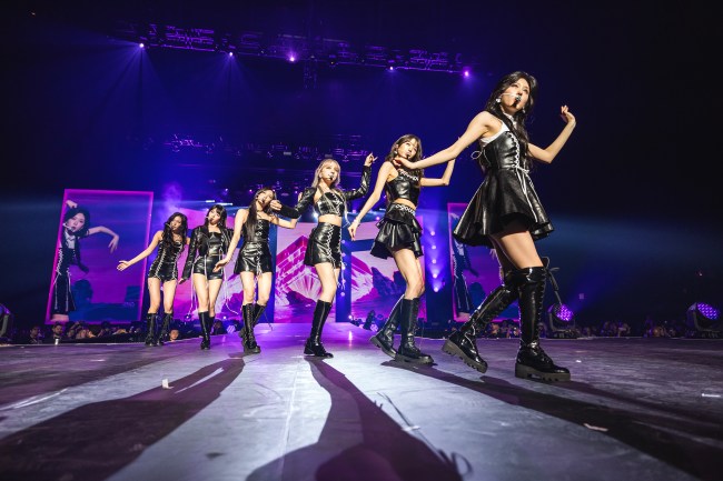 K-Pop Girl Group IVE, six Asian women ANYUJIN, GAEUL, REI, JANGWONYOUNG, LIZ, and LEESEO, dancing with black leather outfits on stage.