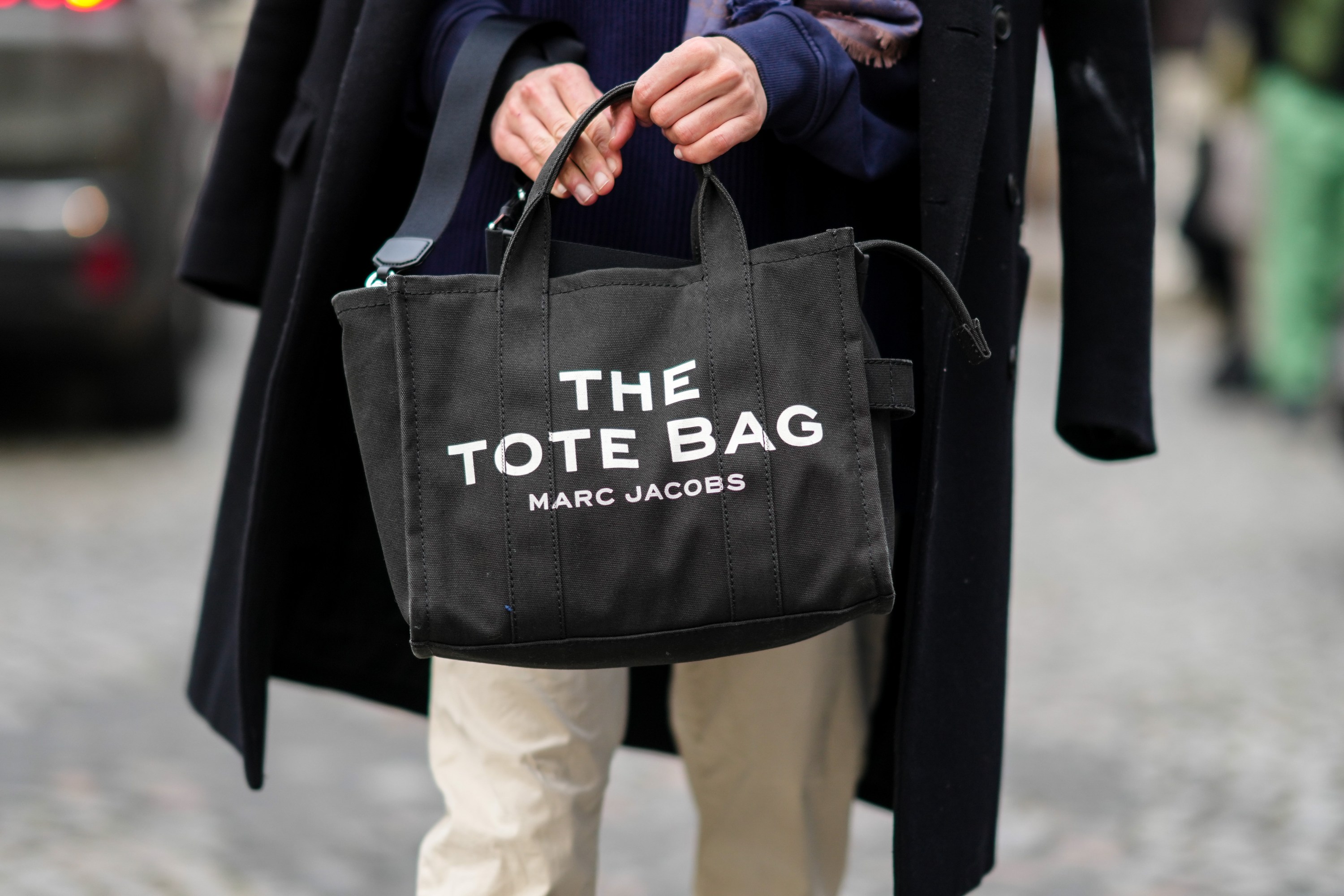 A guest wears a navy blue ribbed wool pullover, a black long coat, a black Tote-bag handbag from Marc Jacobs, beige pants, outside Hermes, during Paris Fashion Week - Menswear F/W 2022-2023, on January 22, 2022 in Paris, France.