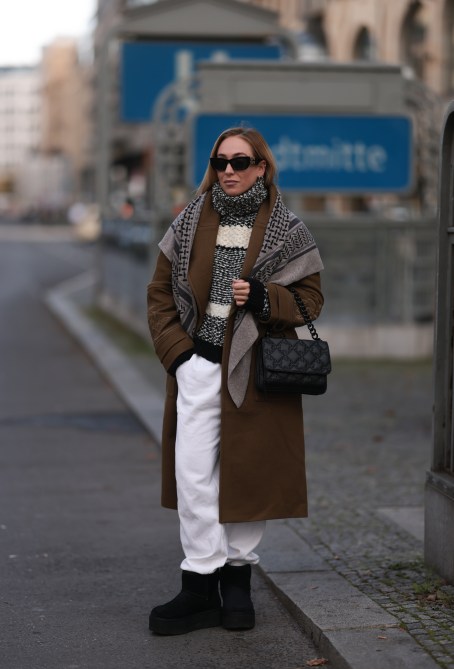 Sonia Lyson wearing white pants, a beige and black turtleneck, a checked scarf, a brown coat, and black Ugg platform boots on October 28, 2022 in Berlin.