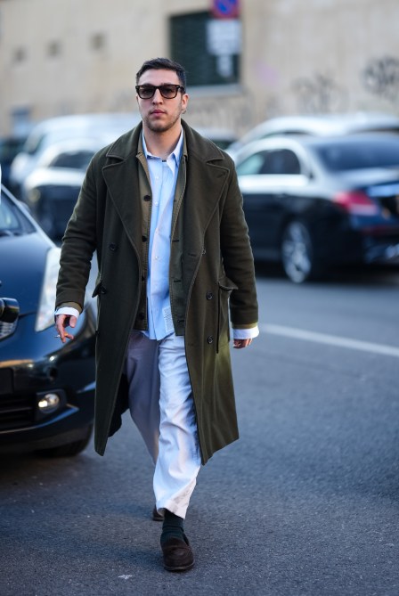 A man wears sunglasses, a green khaki long coat, a pale blue shirt, white wide-leg pants, wool socks, brown suede shoes,