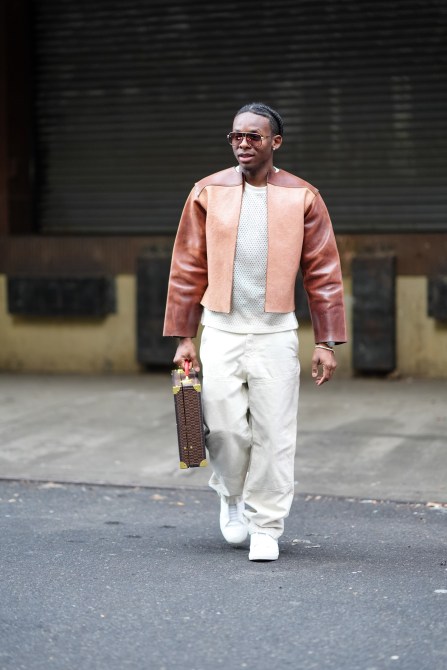 A man wears sunglasses, a beige and brown leather jacket , a white mesh top, white denim jeans pants , white sneakers shoes