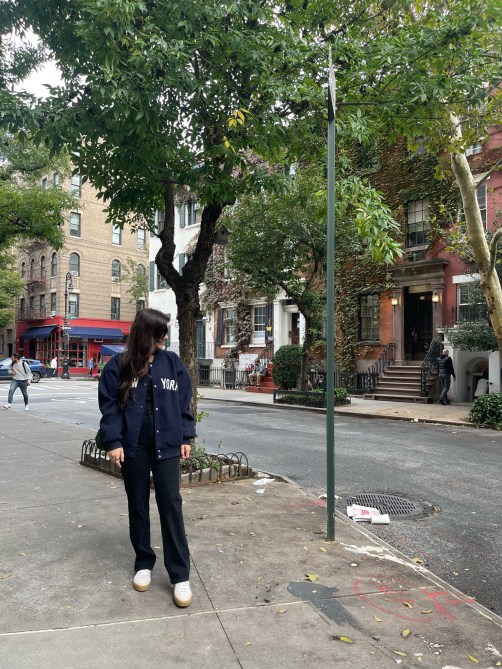 A woman standing on a New York City street in black pants, white shoes, and a navy blue jacket that says "New York."