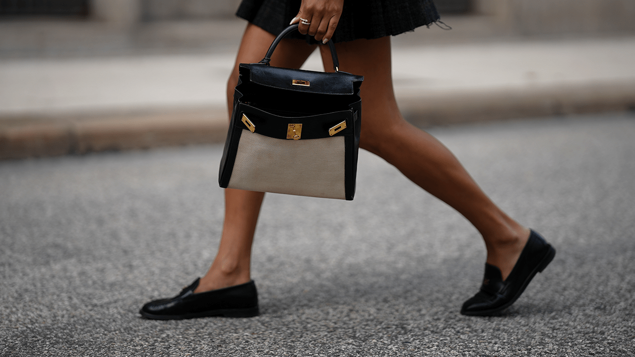 Cherifa Akili is seen wearing a black Noeh blouson in satin look from Someday; an ecru lightweight ribbed t-shirt from Arket; a black pleated mini skirt with tartan check pattern and raw hem from Miu Miu; a pair of shiny black calfskin leather moccasins with thin sole and small golden logo details from Chanel; a black leather Kelly 28 bag with golden hardware and beige canvas front from Hermès; a yellow gold Hélice spiral bracelet from ÉLOUISE; a yellow gold Toi et Moi ring stacked with diamonds from ÉLOUISE on June 4, 2025 in Hamburg, Germany.