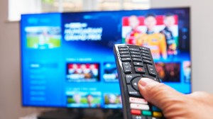 A finger pressing a button on a TV remote with a blurry TV in the background